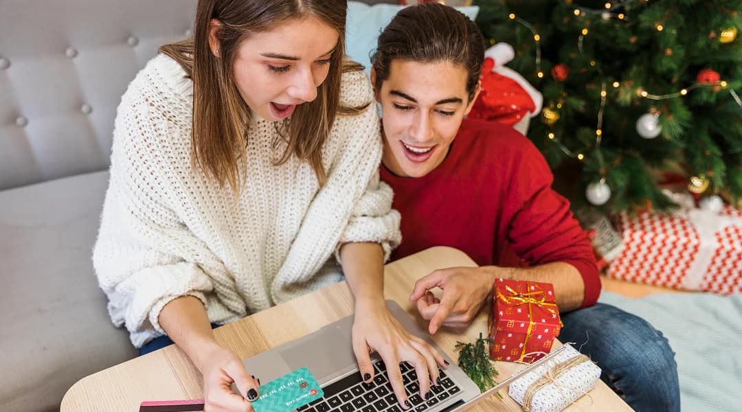 Une jeune femme et un jeune homme devant un ordinateur portable, près d'un sapin de Noël décoré et de cadeaux, préparent leurs achats des fêtes.