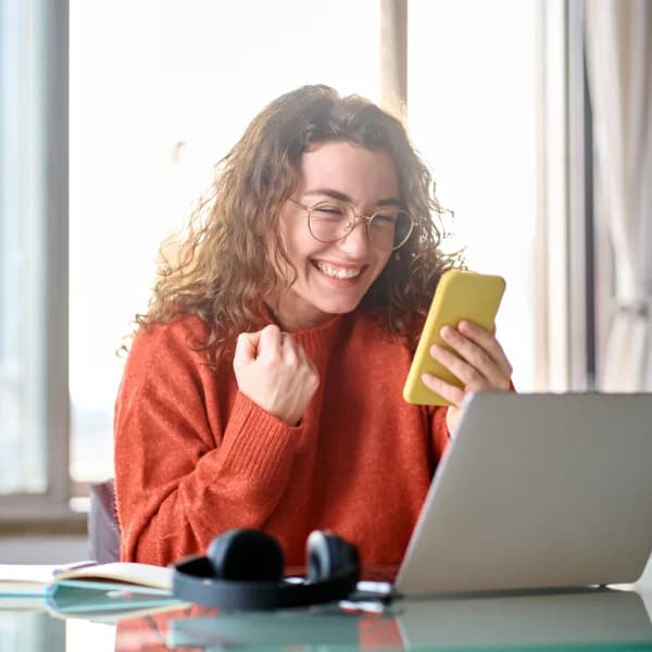 Une femme souriante utilise son téléphone et son ordinateur portable, illustrant la joie de la technologie moderne.