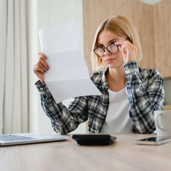 A woman wearing glasses holds a document, looking carefully, probably related to her credit score.